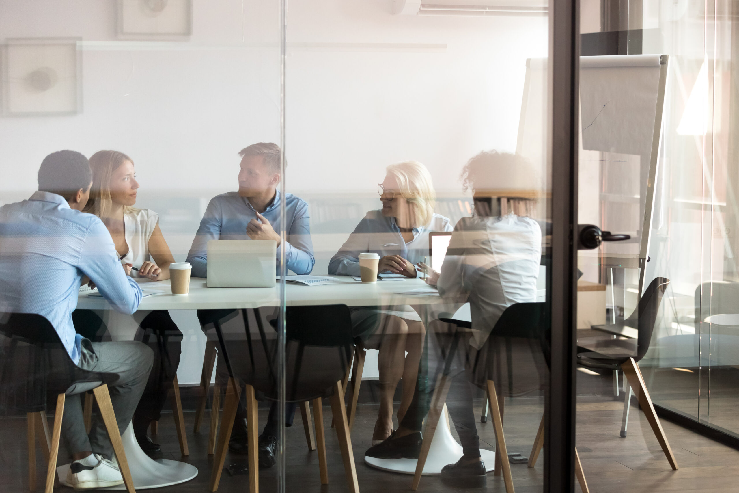 Advertising department brainstorming at modern office boardroom behind closed doors, view through the glass wall. Diverse staff led by ceo discussing new project sharing ideas thoughts and sales pitch